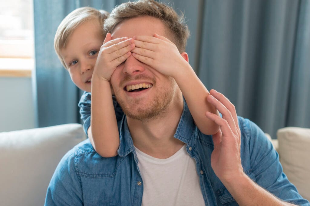 New Dads Need Your Help. Dad sitted with child standing behind and covering dad's eyes with both hands.